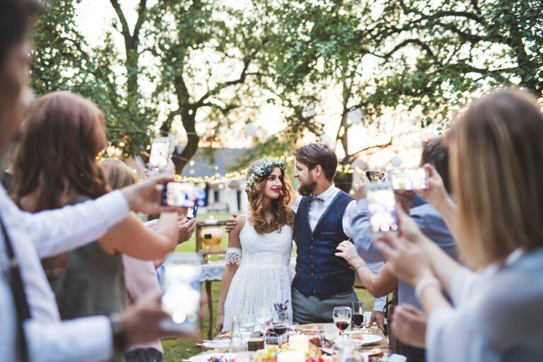 guests-with-smartphones-taking-photo-of-bride-and-groom-at-wedding-reception-outside-